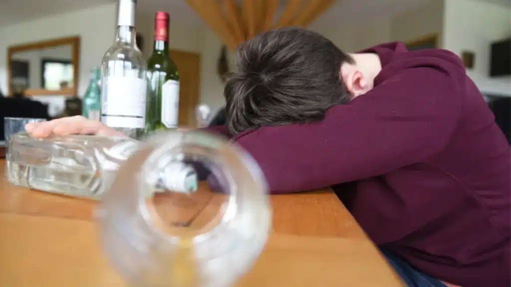 Alcoholism in Teens: Is My Teen an Alcoholic? 5 Teen boy with his head on a table, surrounded by alcohol bottles. Text explains the dangers of drinking alcohol at a young age