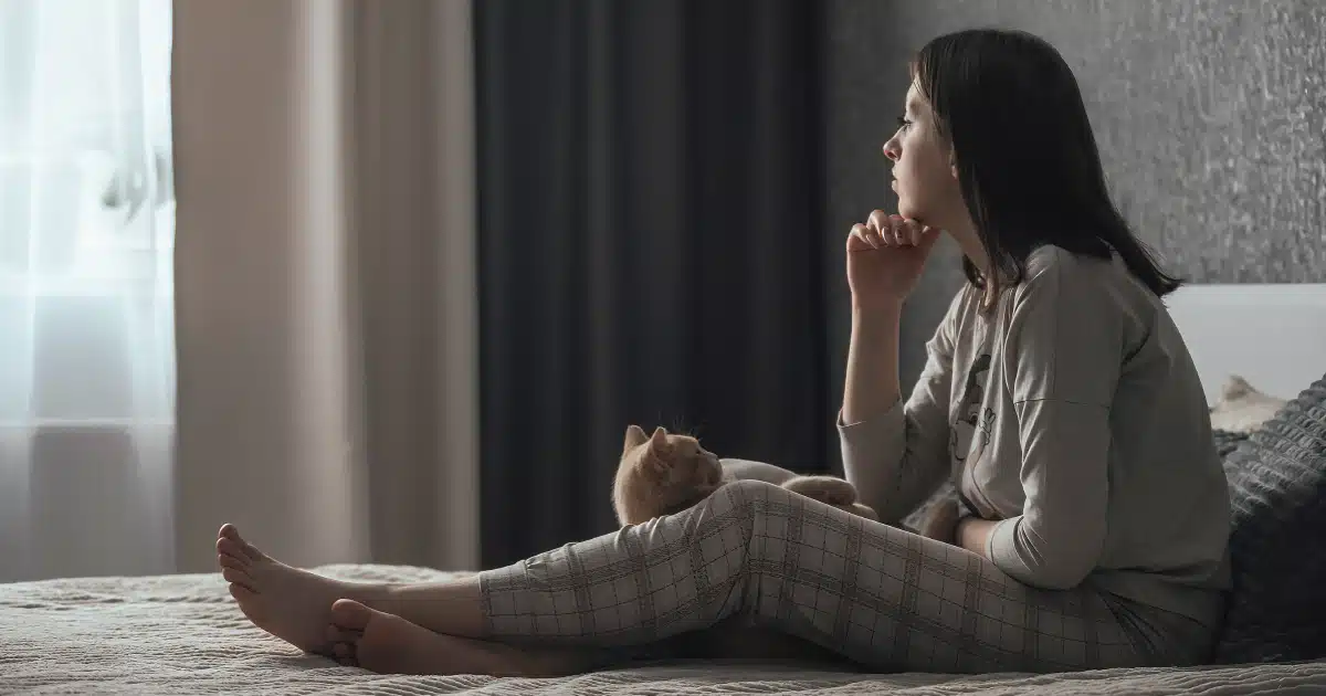 Woman sitting on a bed in a dark room, looking out a window.