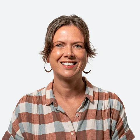 Portrait of a woman with short brown hair wearing hoop earrings and a plaid shirt, smiling against a light gray background.