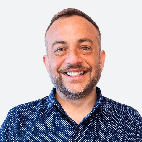 Professional headshot of a man with short hair and a beard wearing a navy blue patterned shirt, smiling against a light background.