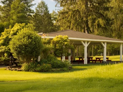 A peaceful outdoor pavilion surrounded by green trees and manicured grass, with picnic tables underneath, on a sunny day.